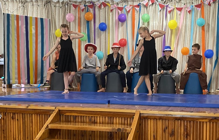 Two girls dancing and six boys drumming on stage with a back drop of ribbons and balloons
